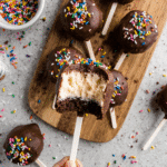 Hand holding a bitten ice cream cake pop showing a vanilla ice cream center and chocolate cake base, coated in chocolate with rainbow sprinkles.